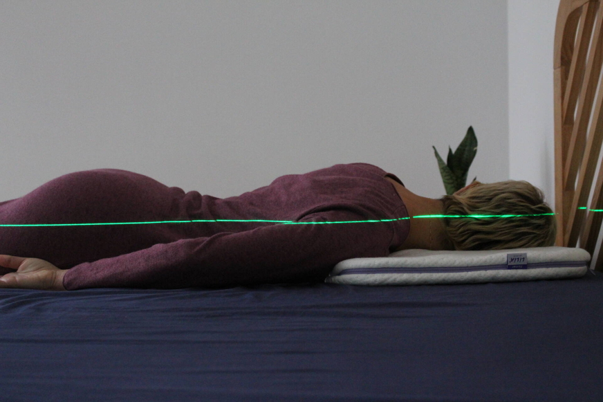 Pessoa de cabelo curto claro deitada de barriga para baixo numa almofada branca com um laser verde a mostrar o alinhamento da coluna.